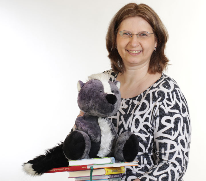 Porträt der Autorin Corinna Wieja mit Bücherstapel, auf dem das Plüsch-Stinktier Teo aus ihren „Teo Dorant“-Kinderbüchern sitzt.
