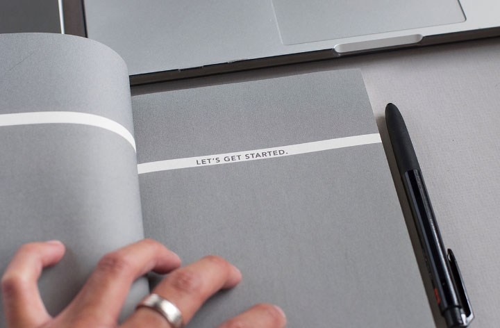 Auf dem Bild ist ein graues Notizbuch mit der Aufschrift Let's get started zu sehen, darauf liegt eine Hand, daneben ein Kugelschreiber