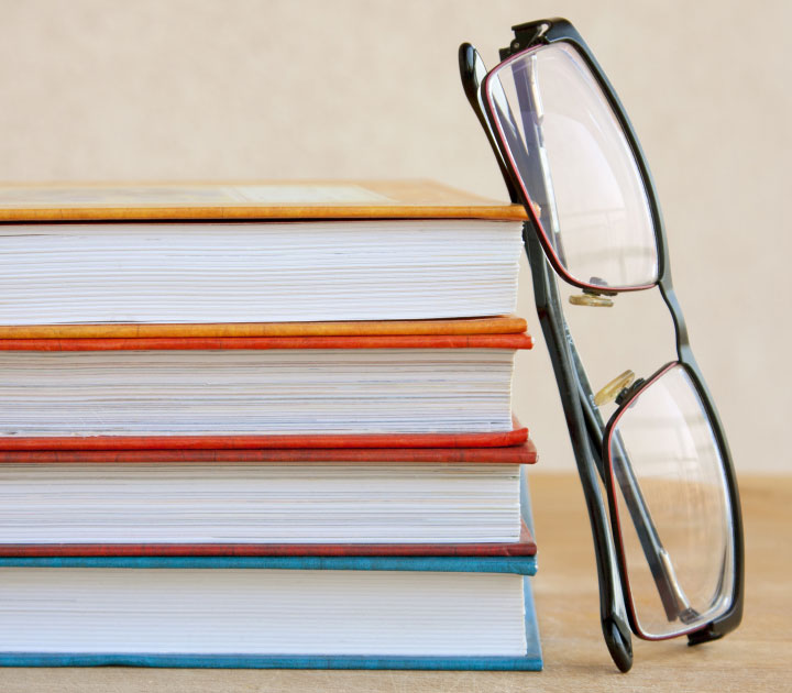 Buchstapel aus jeweils einem blauen, gelben, roten und orangen Buch, an den eine Brille gelehnt ist